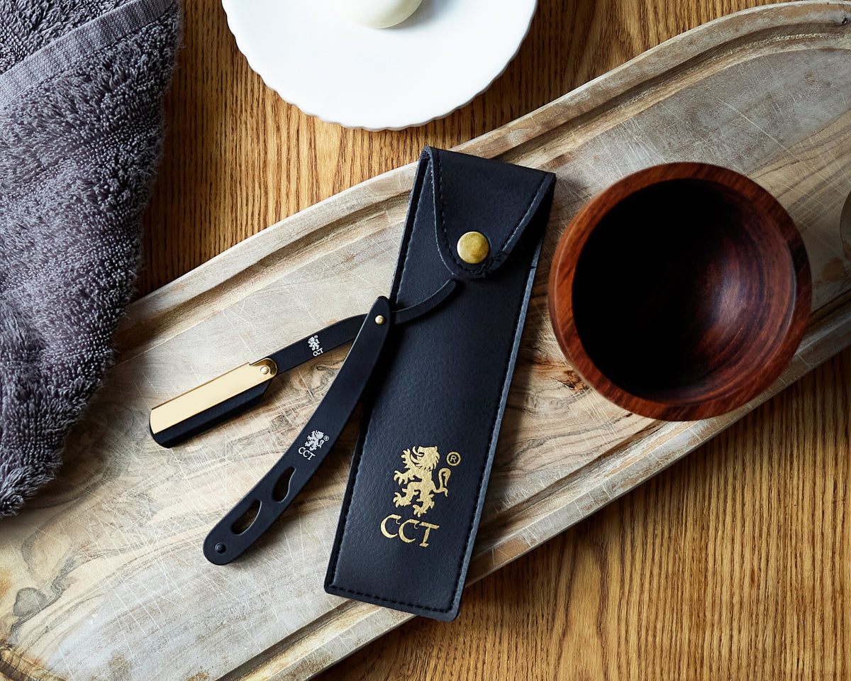 A black and gold cutthroat straight razor with a carry pouch and a wooden shaving cream bowl on a wooden board